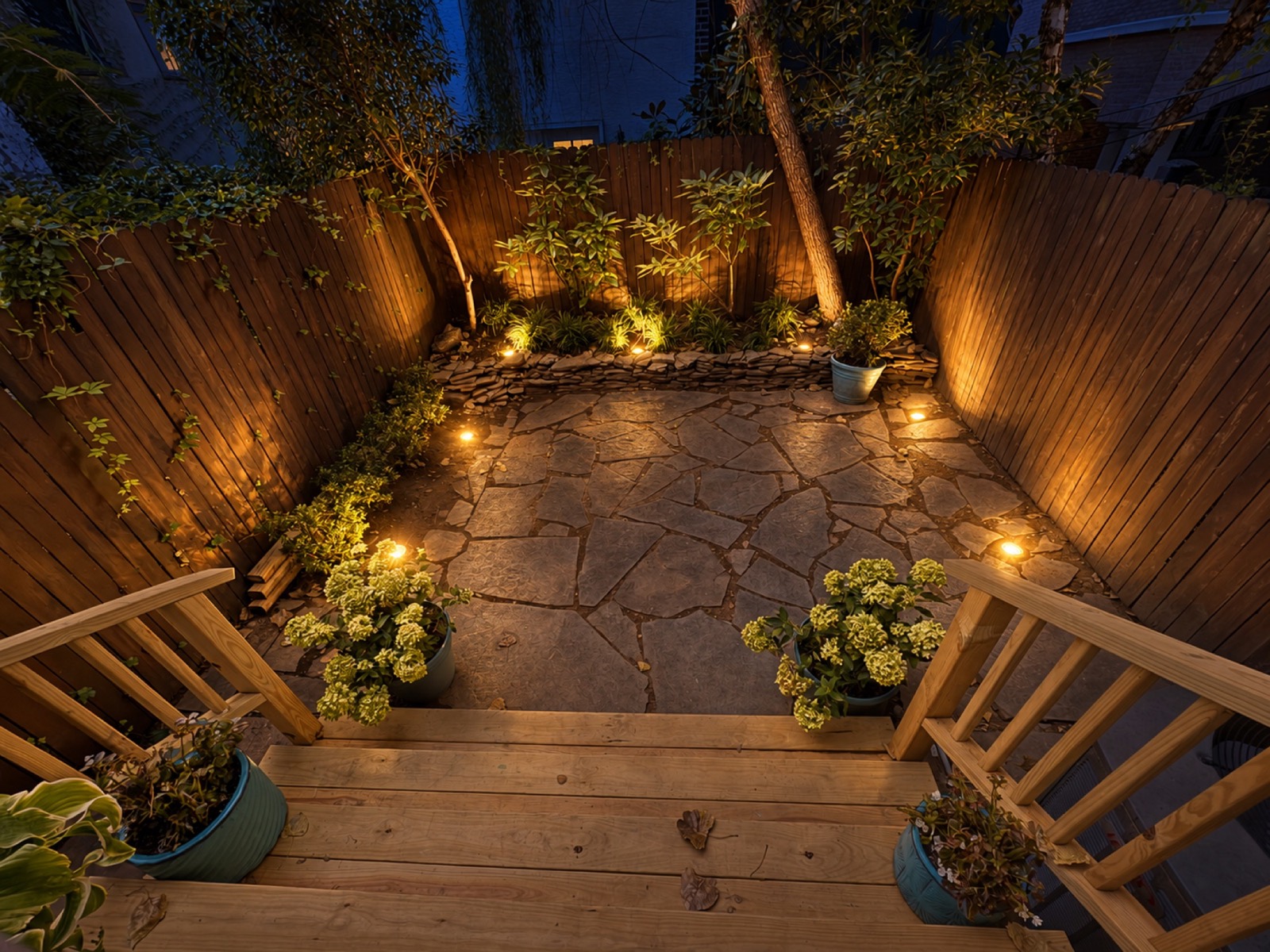 Queen Village backyard at night with warm ground lighting illuminating the stone patio, planting, and fence